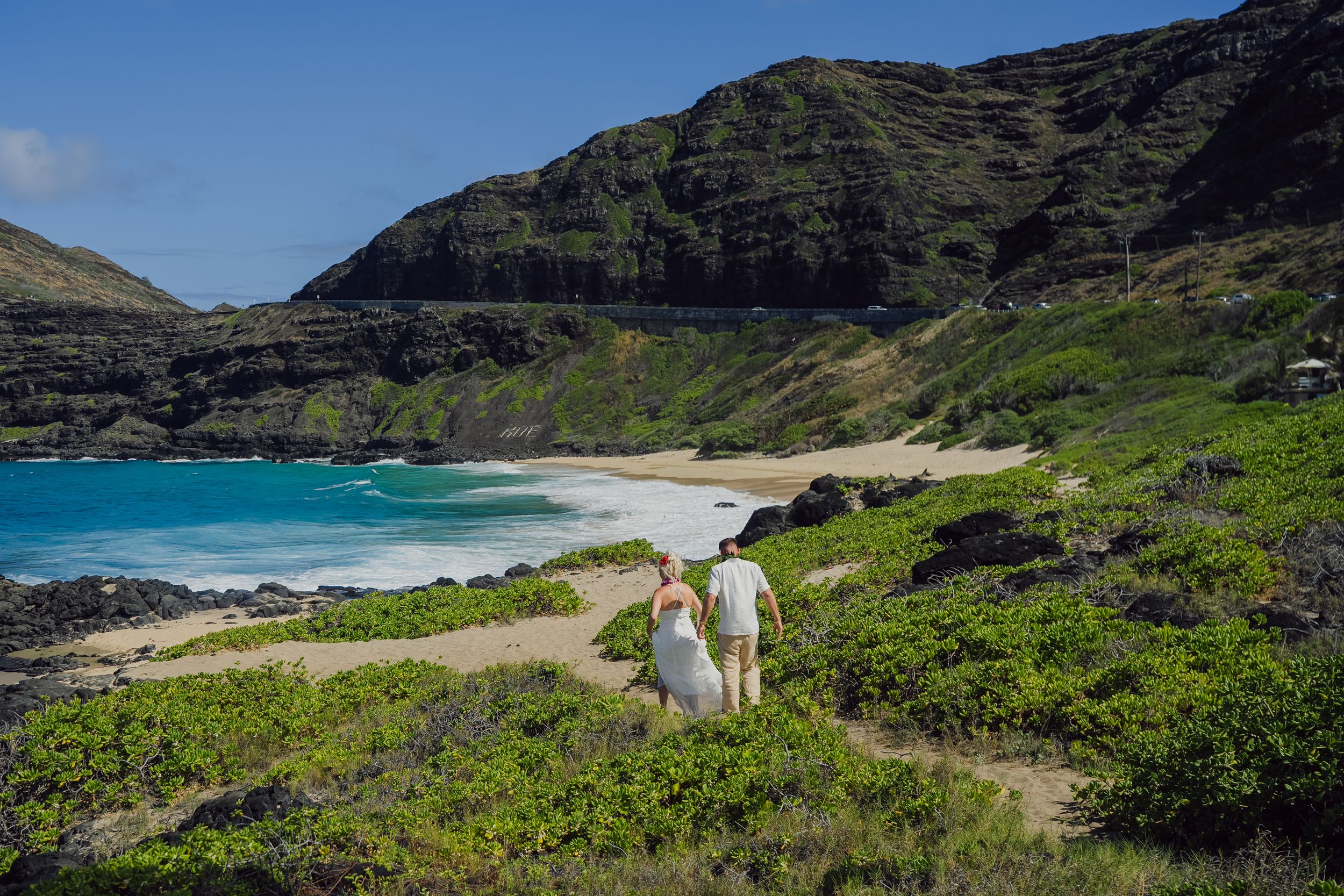 Couple in white walking hand-in-hand along a scenic Hawaii beach trail with turquoise water and lush green mountains.