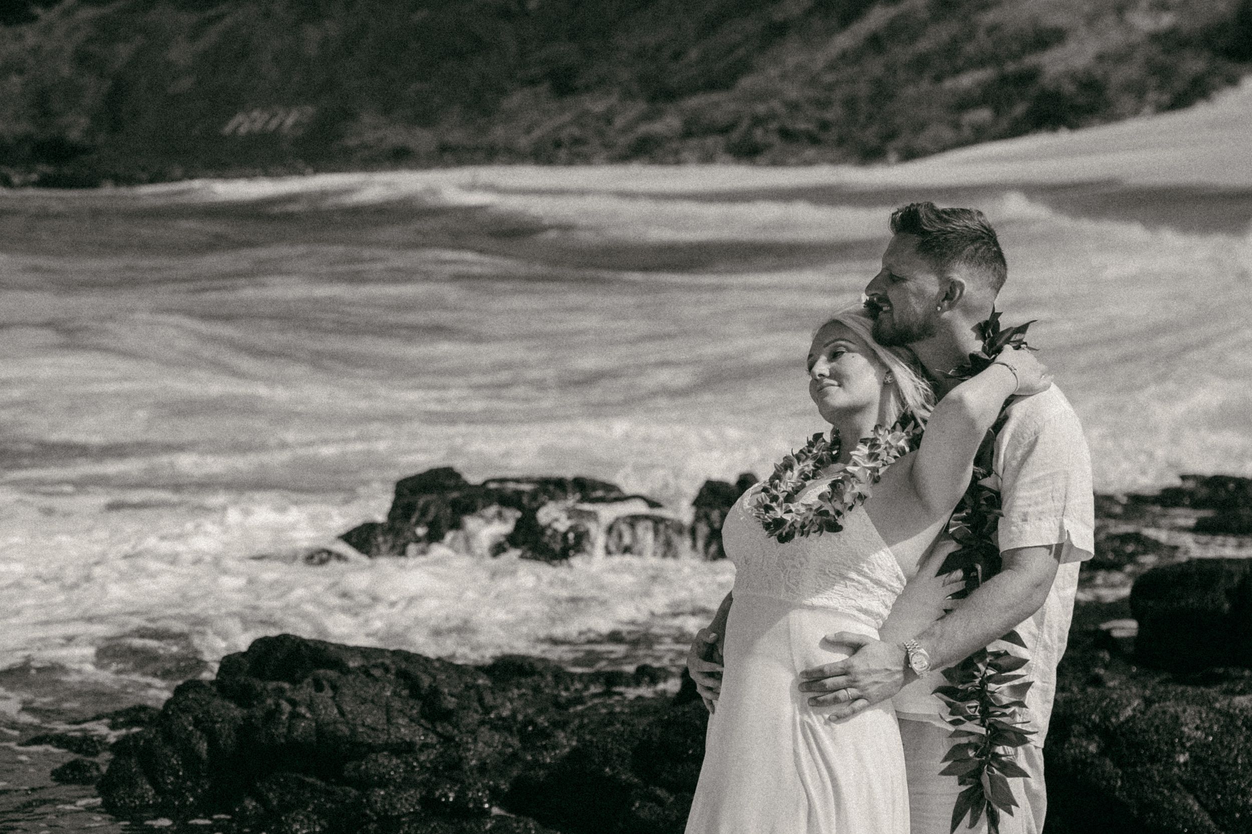 Black and white photo of a couple embracing on rocky Hawaiian shore, woman wearing floral lei, waves crashing behind them.