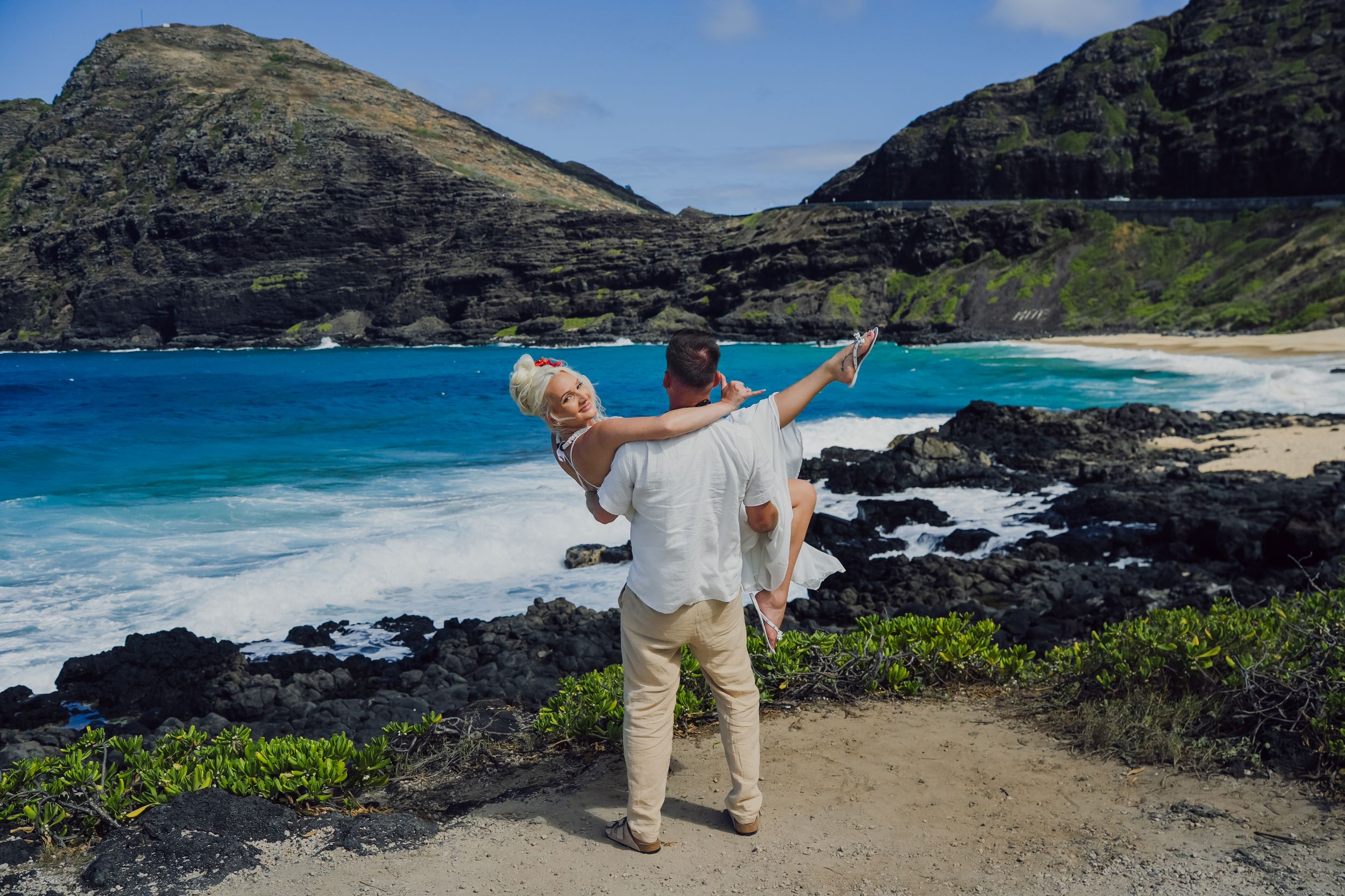Couple embracing on rocky Hawaiian coastline with turquoise ocean waves and lush green mountains in background.