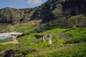 Two brides in white dresses hold hands on a lush green hillside near a Hawaiian beach with dramatic mountains behind.