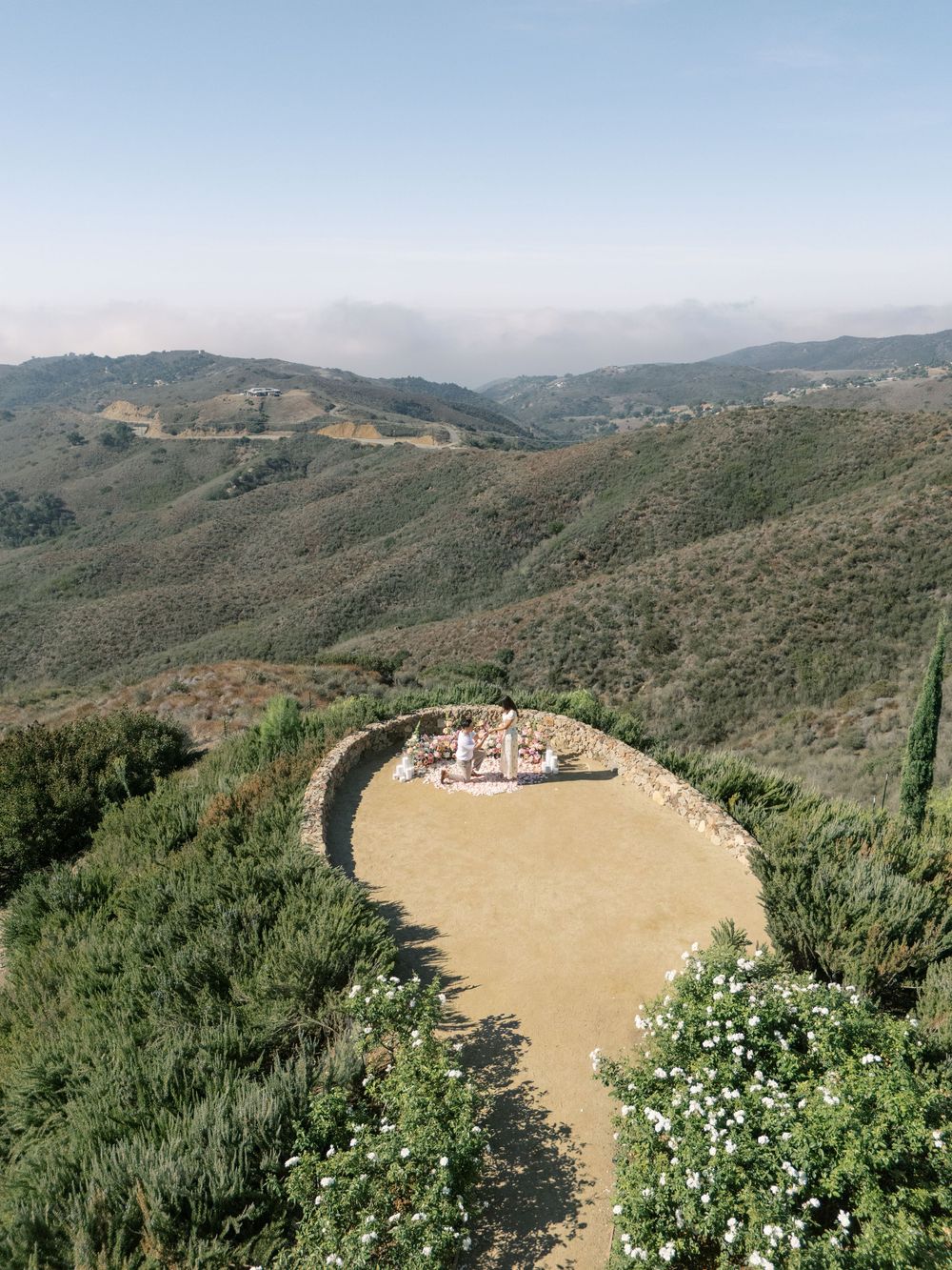 Aerial view of a circular stone ceremony space surrounded by lush green hills and mountain landscape under a hazy sky.