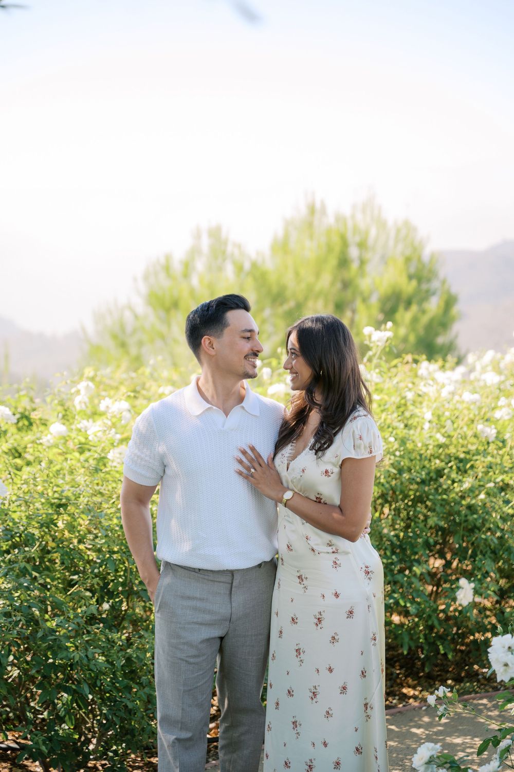 A smiling couple stands together in a sunlit garden with white flowers and green hills, the woman wearing a floral dress.
