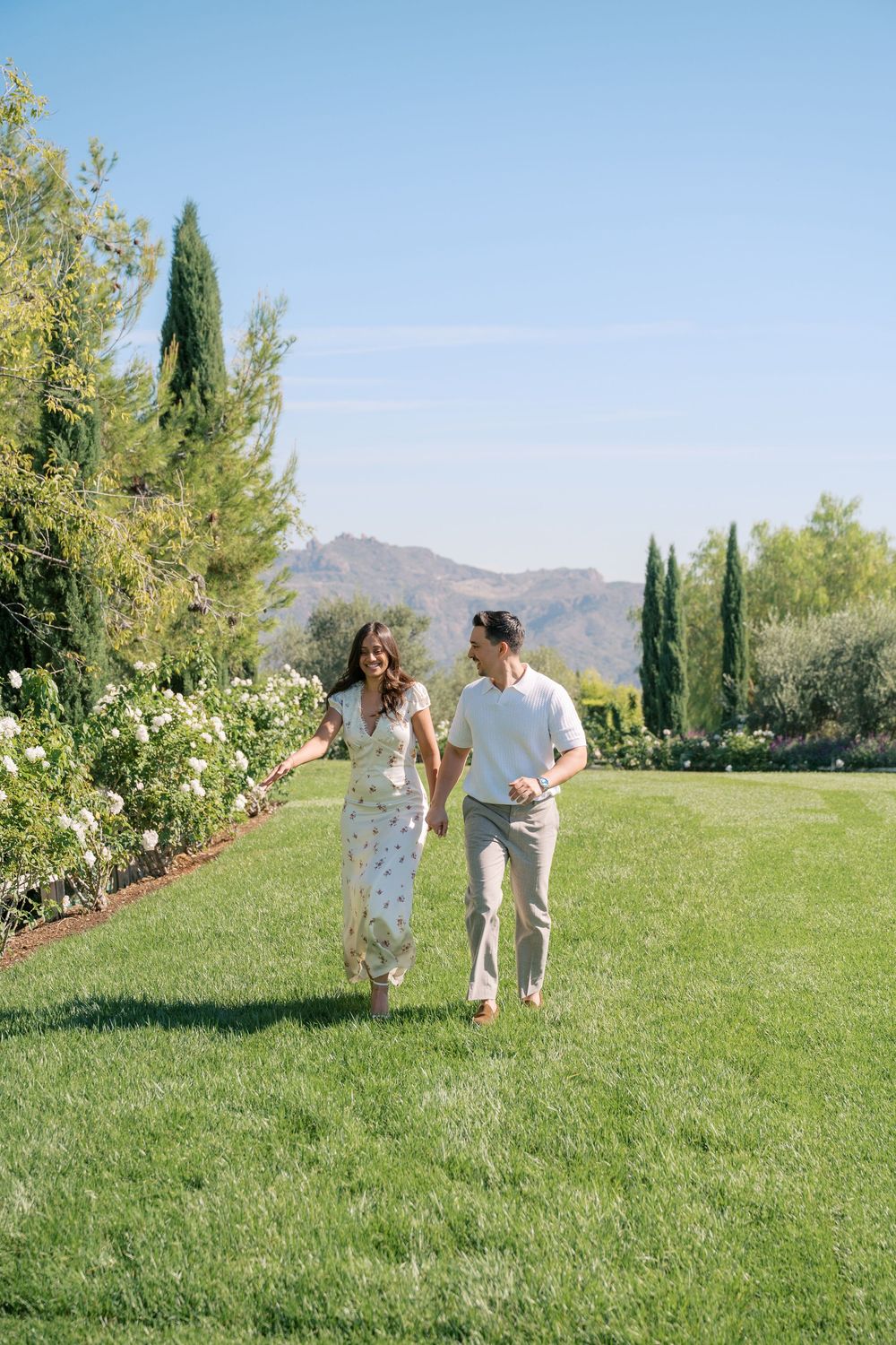 Happy couple holding hands walking across green lawn with cypress trees and mountain views behind them.