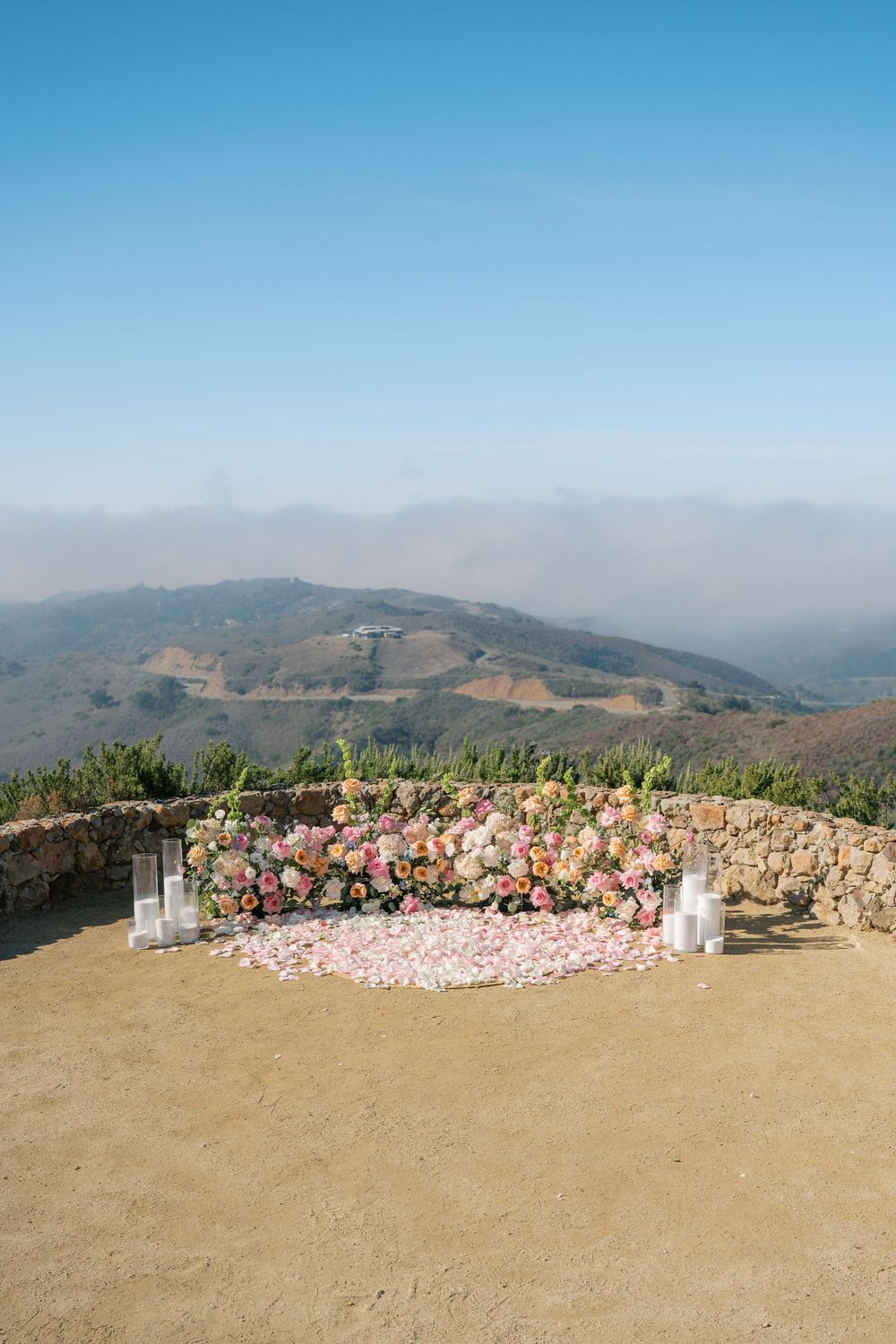 Romantic outdoor proposal setup with colorful floral wall and rose petals on hilltop overlooking misty mountains.