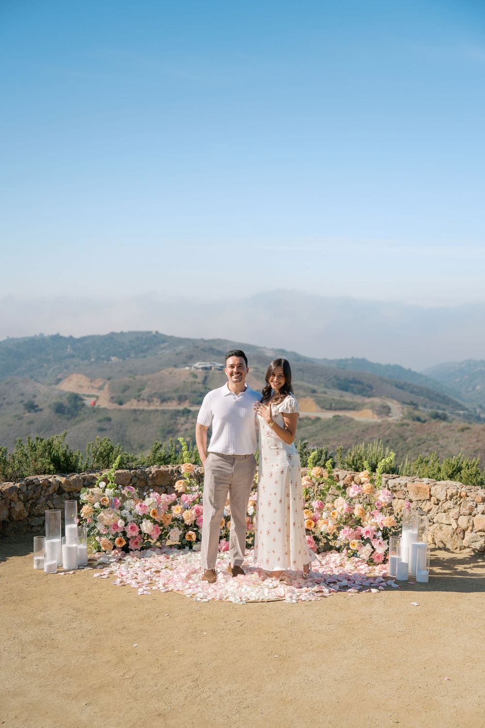 Couple smiling at outdoor proposal setup with pink flowers, rose petals, and candles against scenic mountain backdrop.