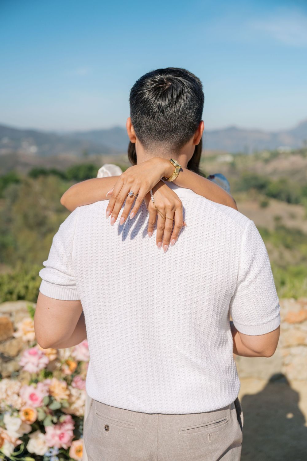 Close-up of woman's hands with engagement ring wrapped around man's shoulders at outdoor proposal site.