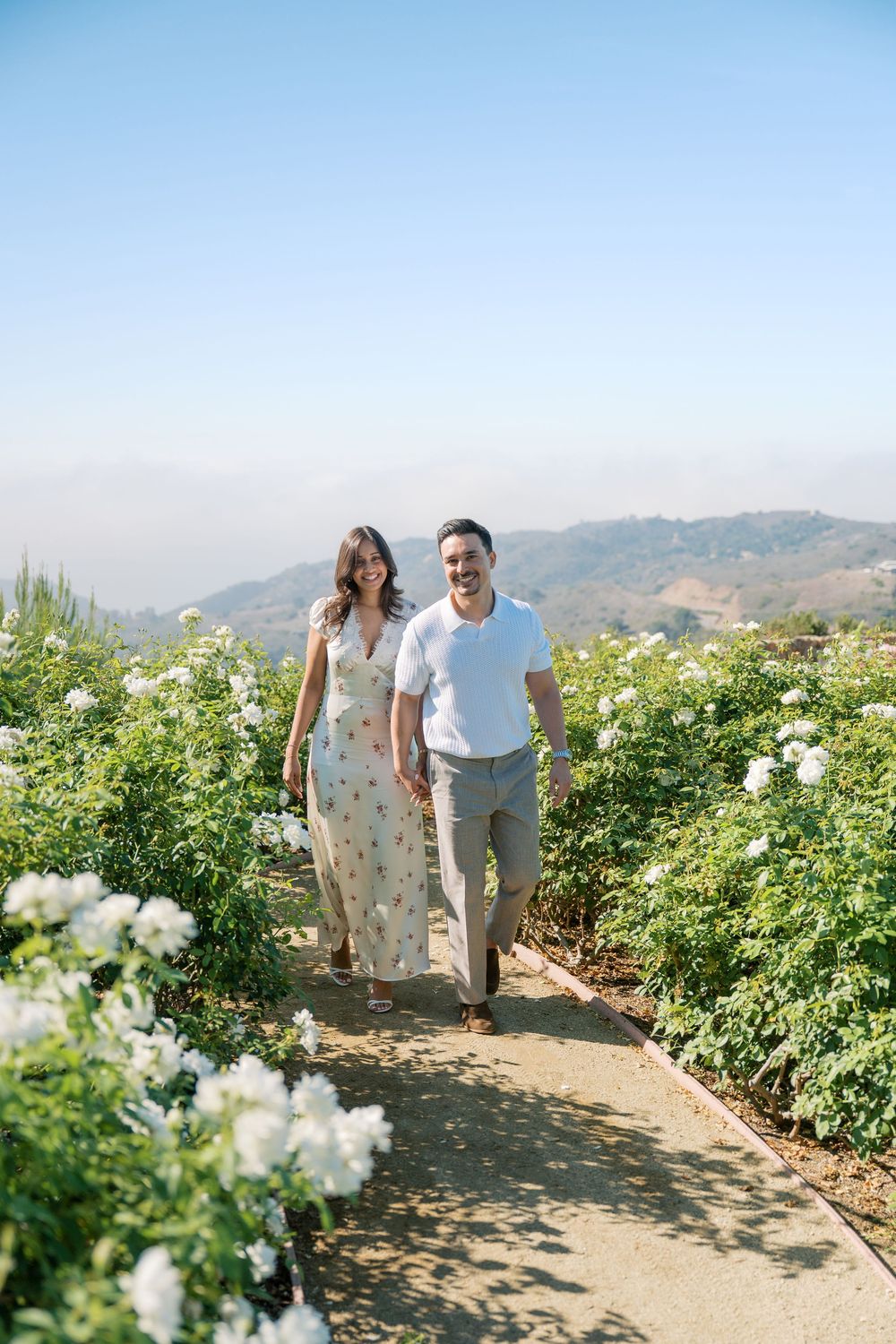 Smiling couple walking hand in hand along garden path lined with white roses and mountain backdrop.