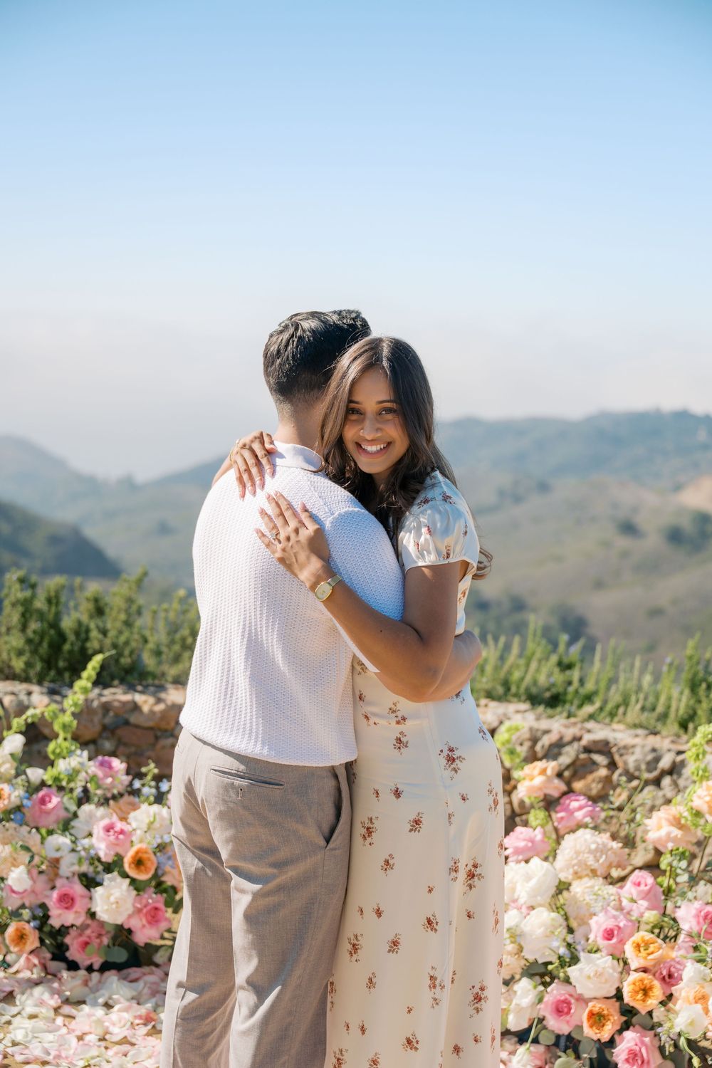 Couple embracing at outdoor proposal spot with colorful floral arrangements and scenic mountain views in background.