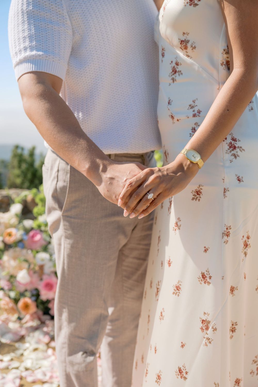 Close-up of newly engaged couple holding hands showing diamond engagement ring with floral backdrop outdoors.