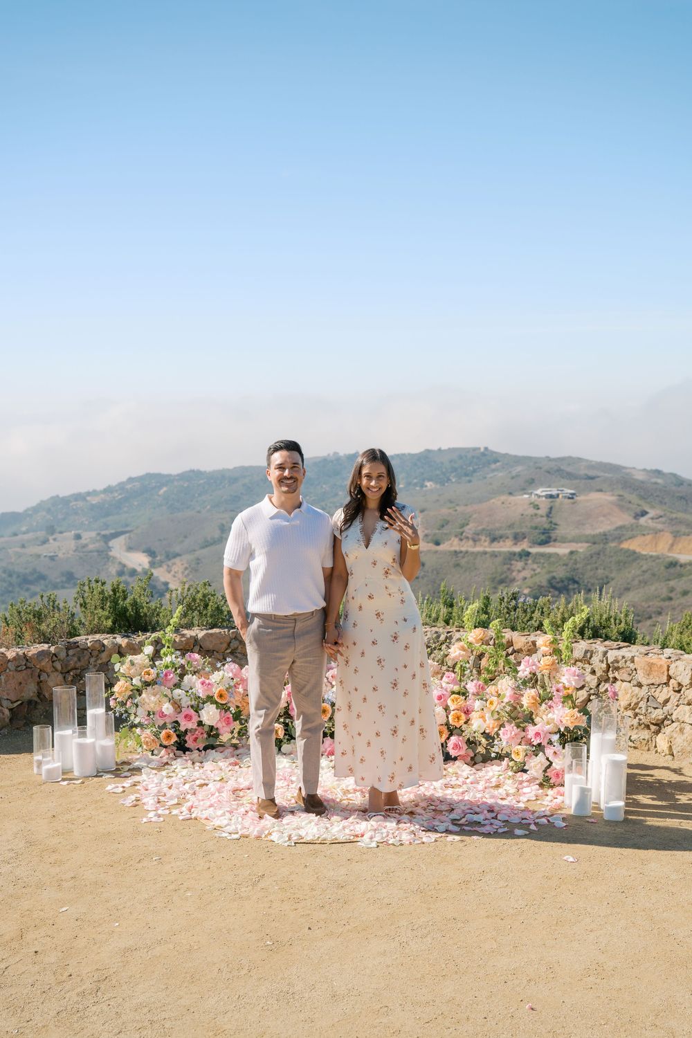 Newly engaged couple smiling and posing before colorful floral backdrop on scenic mountain overlook.