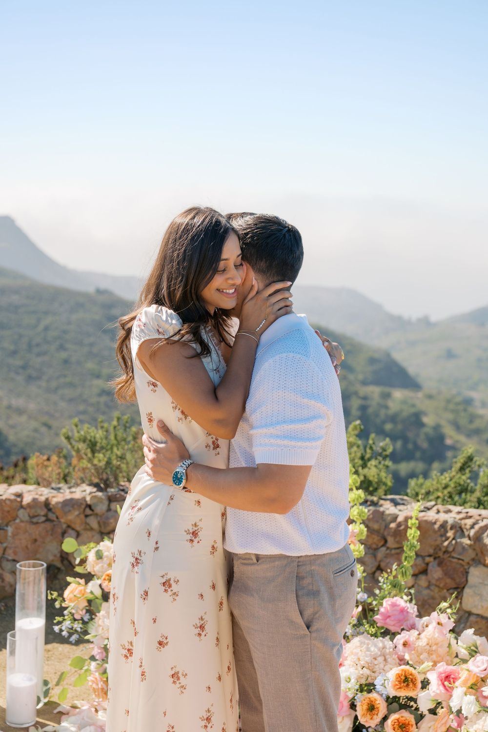 Couple embracing and smiling during outdoor proposal with floral arrangements and mountain views in background.