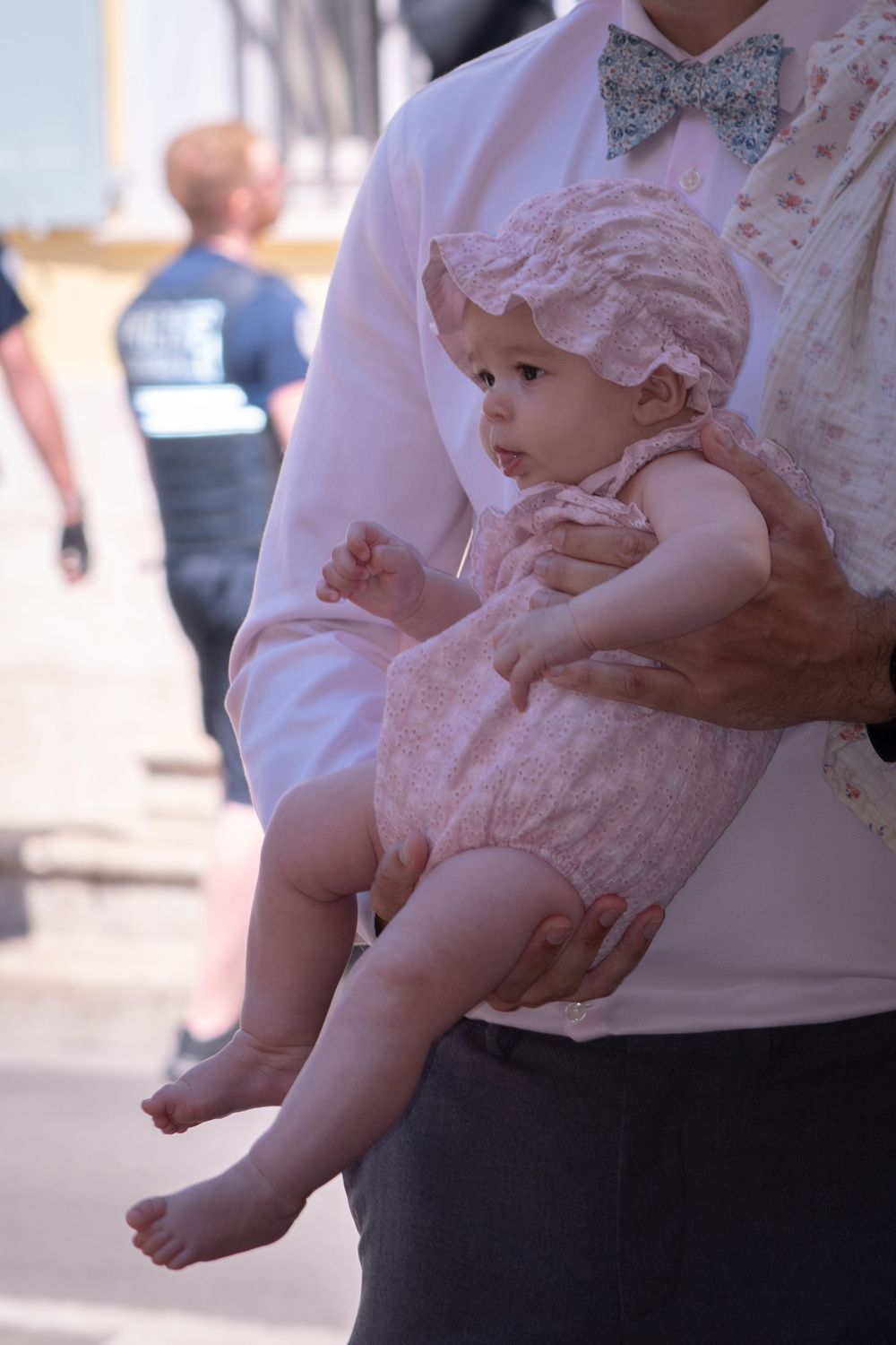 A baby in a pink lace outfit and bonnet being held by an adult in a white shirt with a bow tie outdoors.