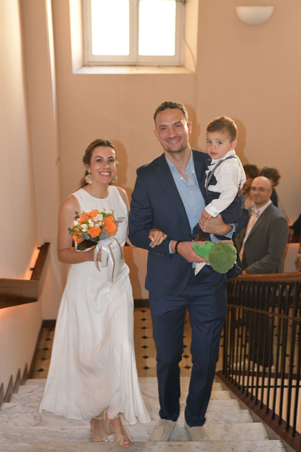 Smiling bride in white dress holding orange bouquet walks with groom in navy suit carrying young child on wedding day.