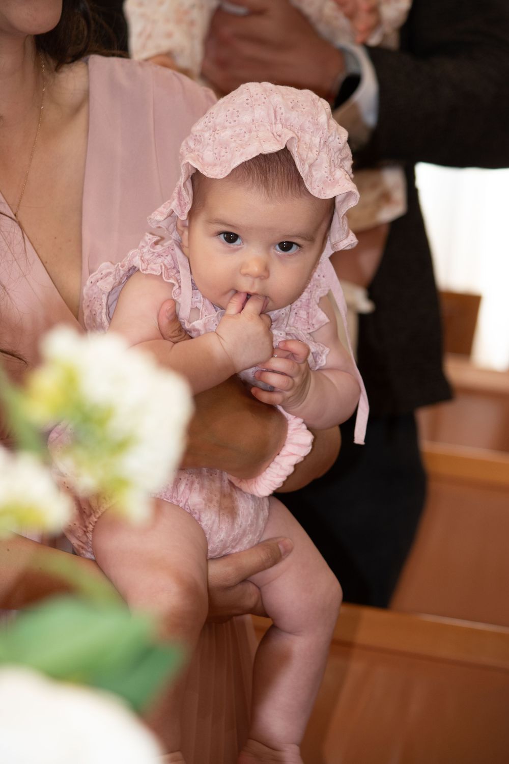 Adorable baby in pink lace bonnet and romper being held during a church baptism ceremony.