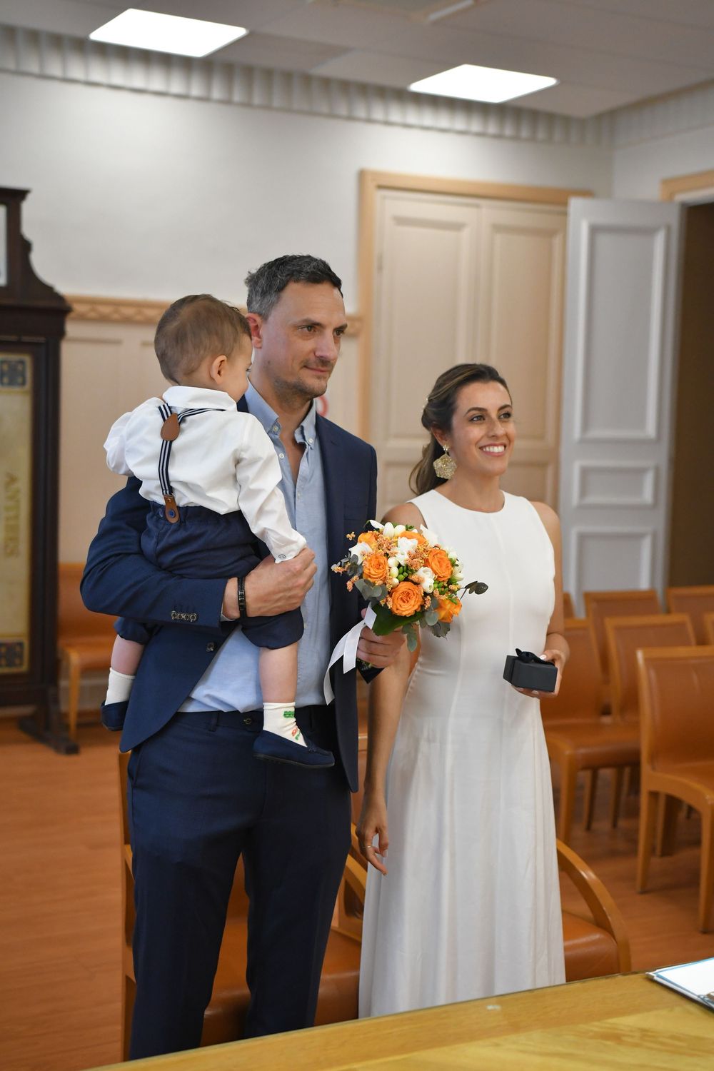 Happy couple in wedding attire holding baby and orange bouquet at civil ceremony indoors.