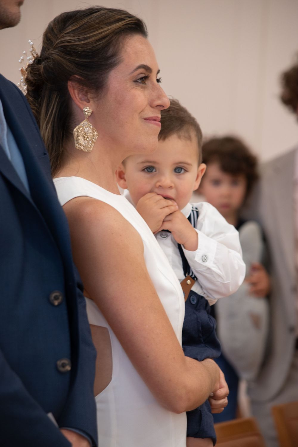 A bride in a white sleeveless dress holds a toddler boy in a suit during an indoor wedding ceremony.