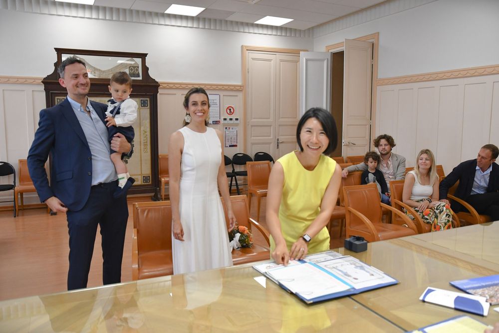 A woman in yellow signs documents at a table during an official ceremony with smiling guests standing nearby in a formal room.