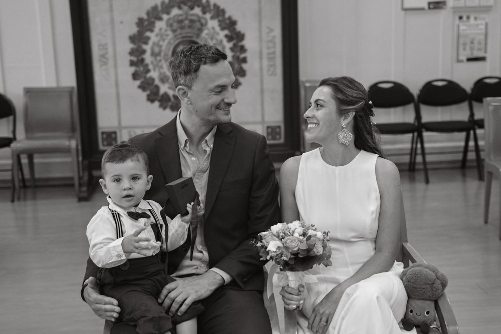 Black and white photo of a newlywed couple sitting with their baby boy, bride holding a bouquet, in a wedding hall.