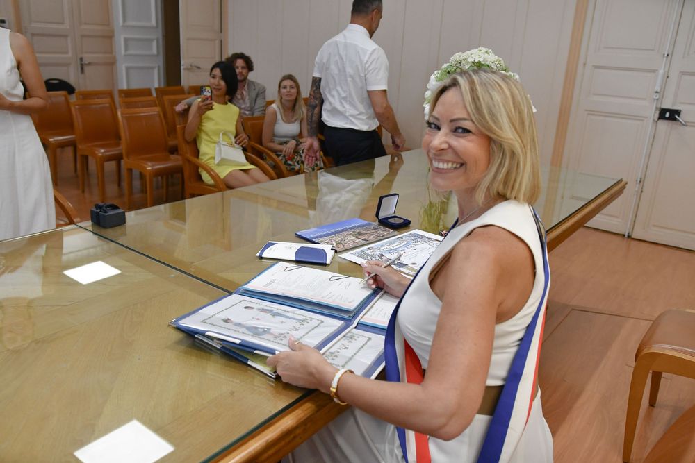 Smiling blonde woman in white dress with tricolor sash at official ceremony table with documents and medals.