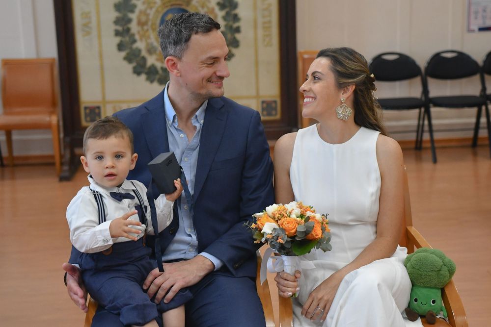 Couple in wedding attire sitting with toddler boy, bride holding orange bouquet, civil ceremony setting.