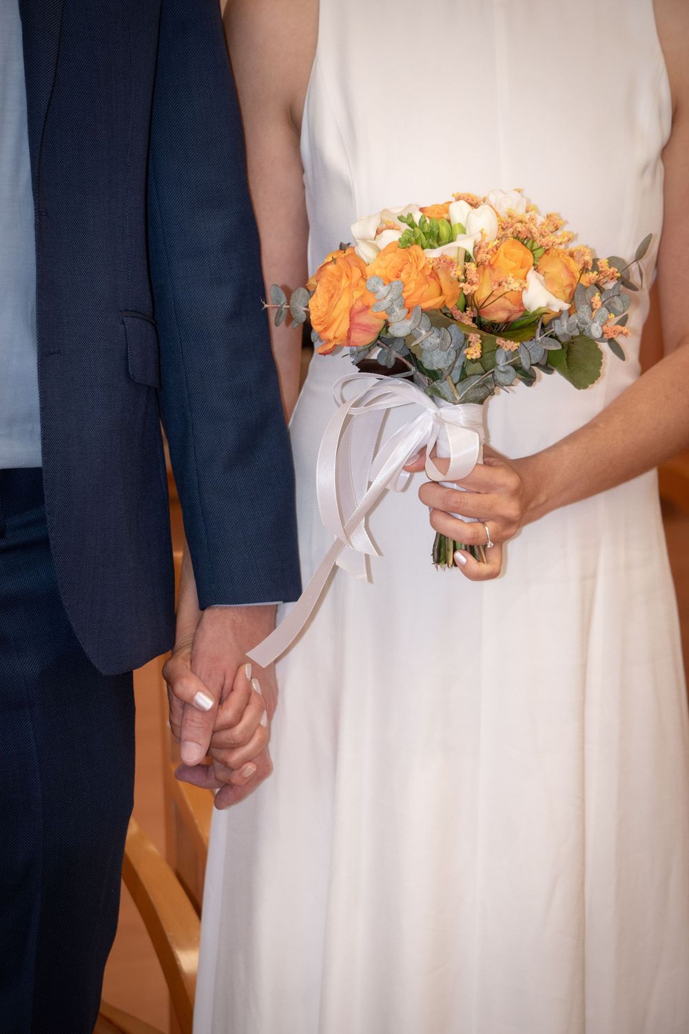 Bride in white dress holding orange rose bouquet with ribbon while holding hands with groom in navy suit at wedding ceremony.