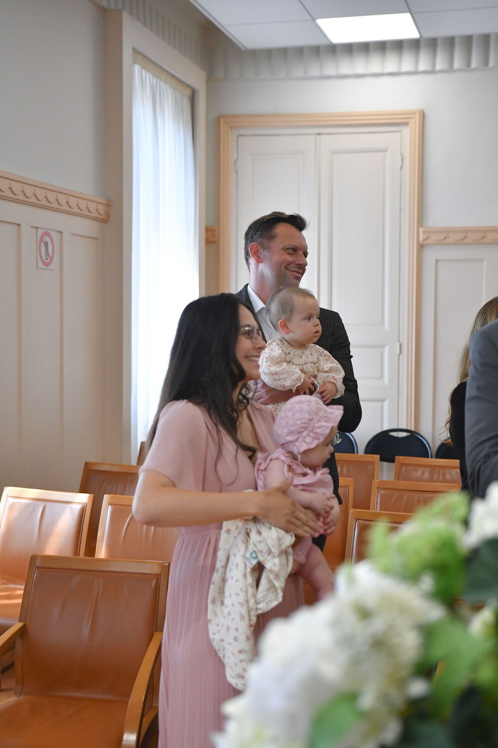 A woman in a pink dress holds a baby at a formal indoor ceremony while a man in a suit smiles behind her.