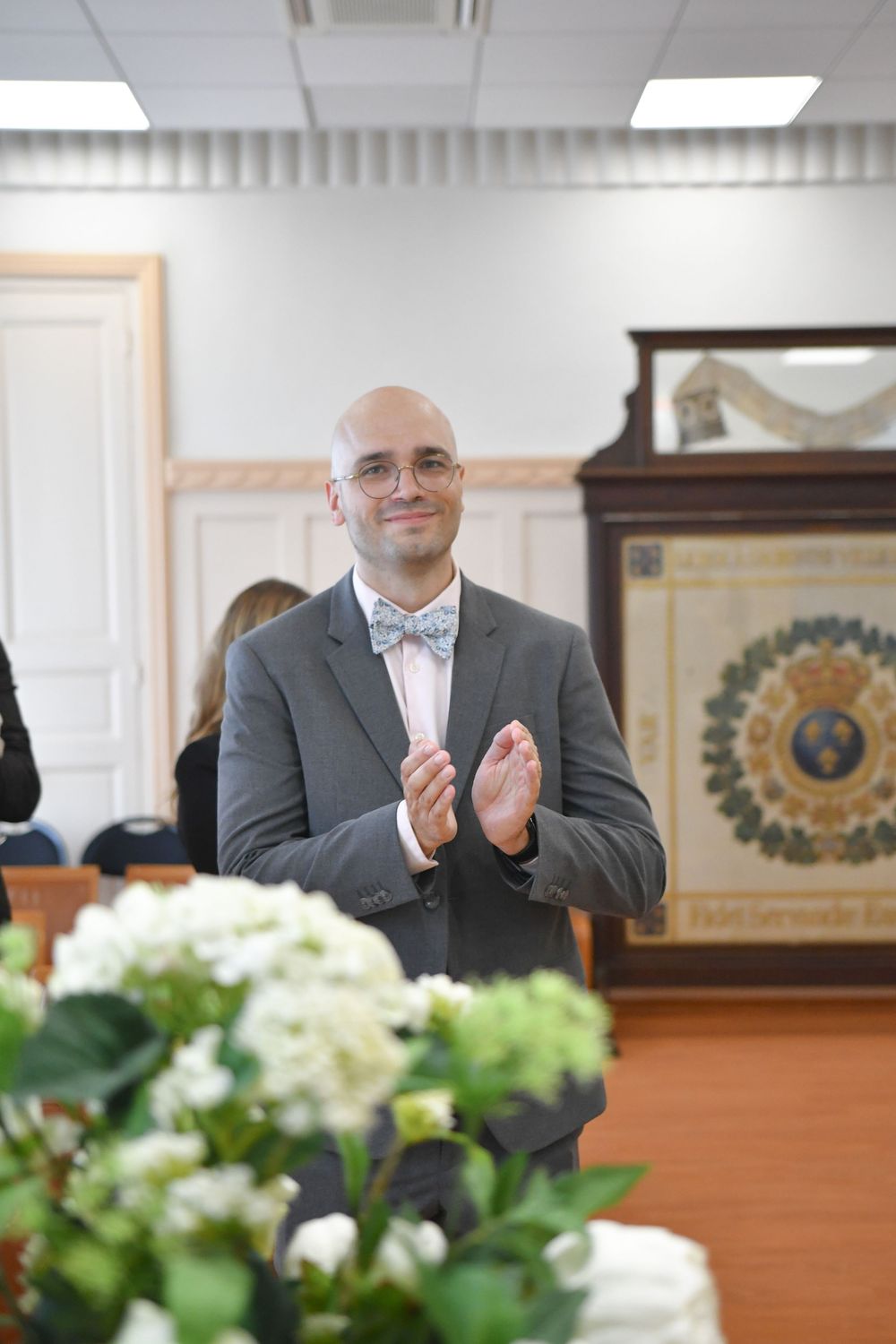 Bald man in gray suit and pink bow tie clapping and smiling at an elegant indoor ceremony with white flowers.