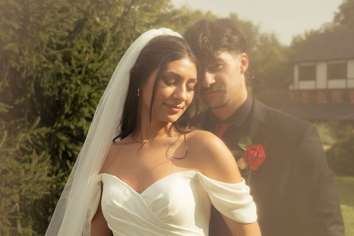 A bride and groom stand in front of an English style cottage. The brides eyes are closed with her groom looks into the camera.