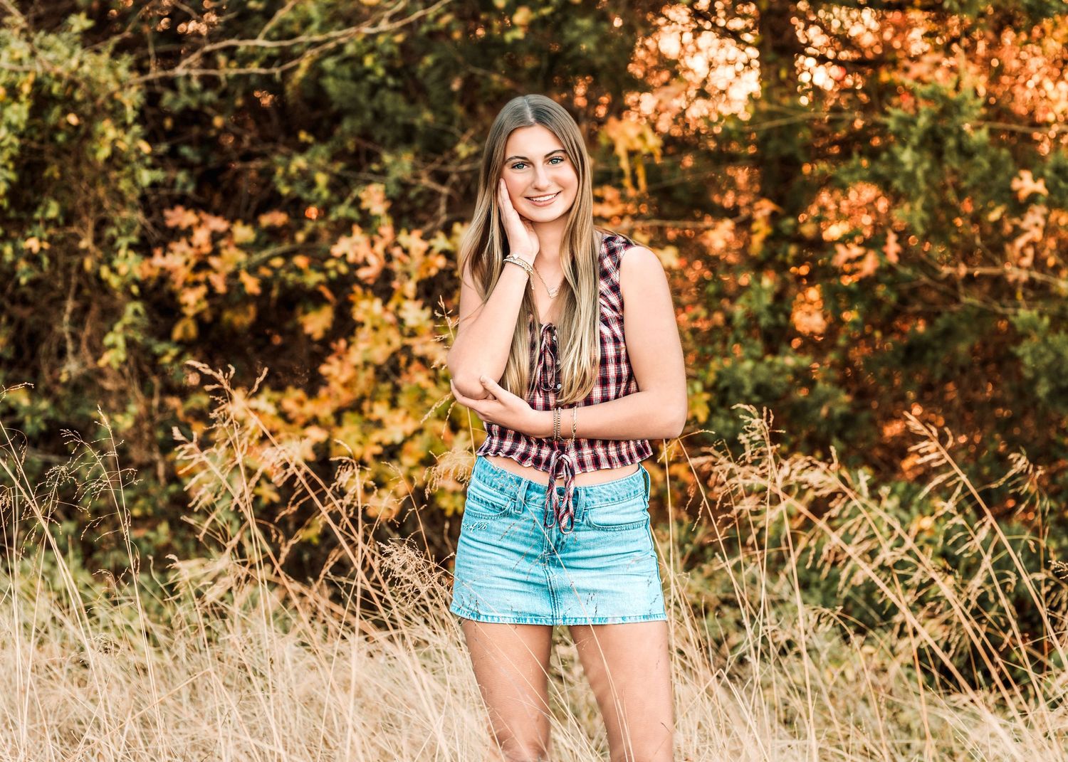 Young woman in plaid shirt and denim skirt smiling in a golden autumn field with colorful fall foliage.