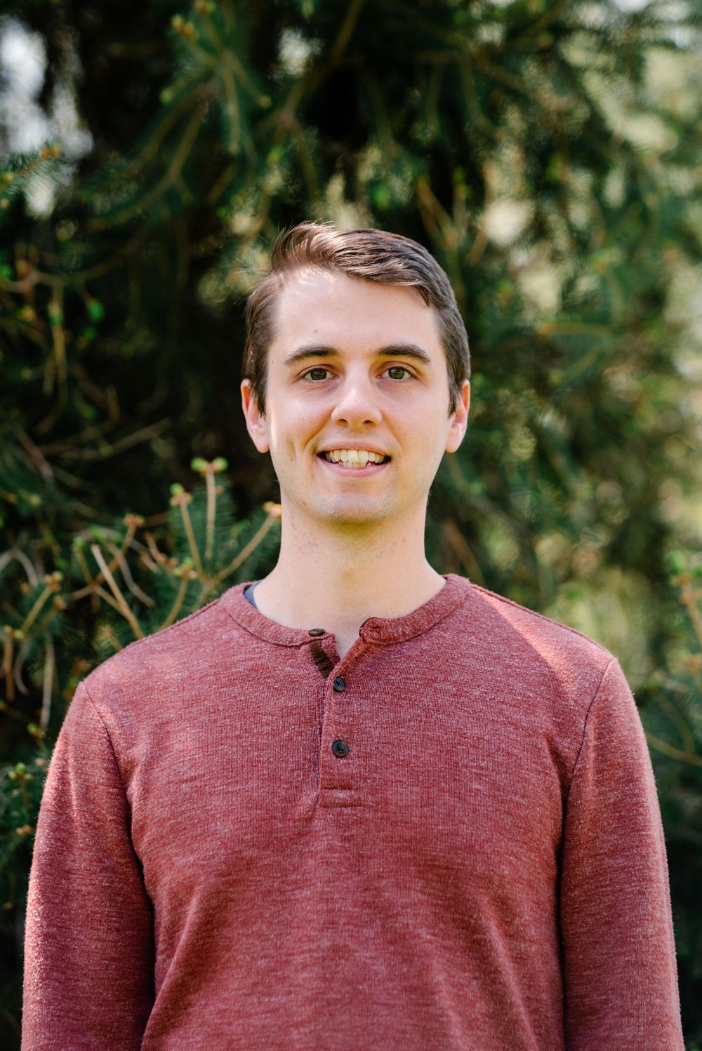 Smiling young man in a red henley shirt standing outdoors in front of green evergreen trees.