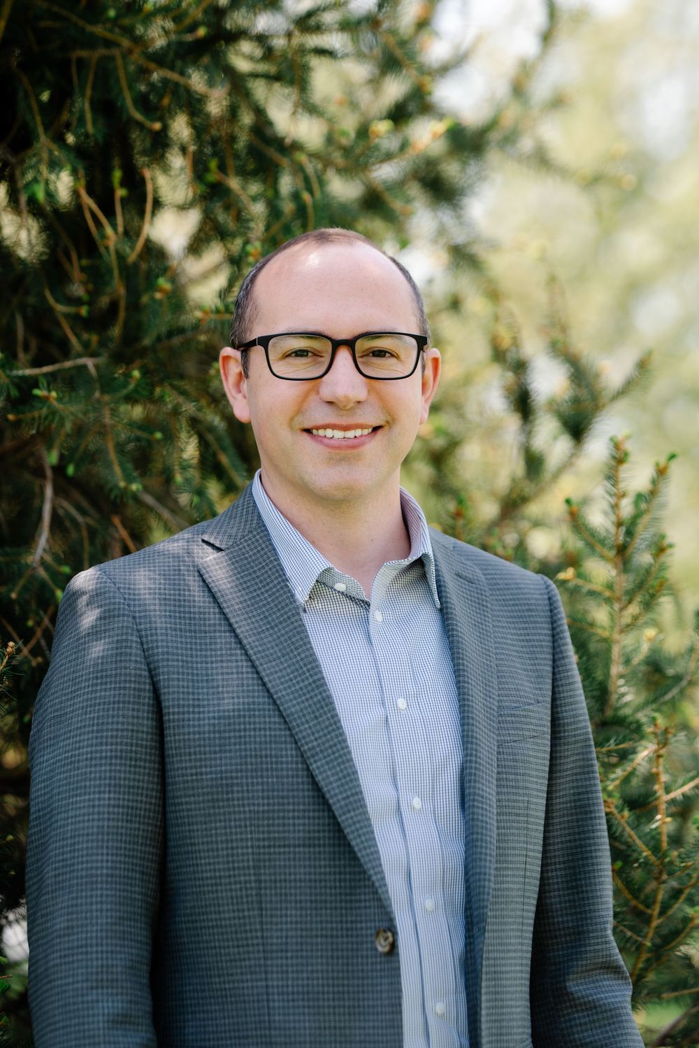 Smiling man with glasses wearing a gray blazer and light blue shirt, standing outdoors near evergreen trees.