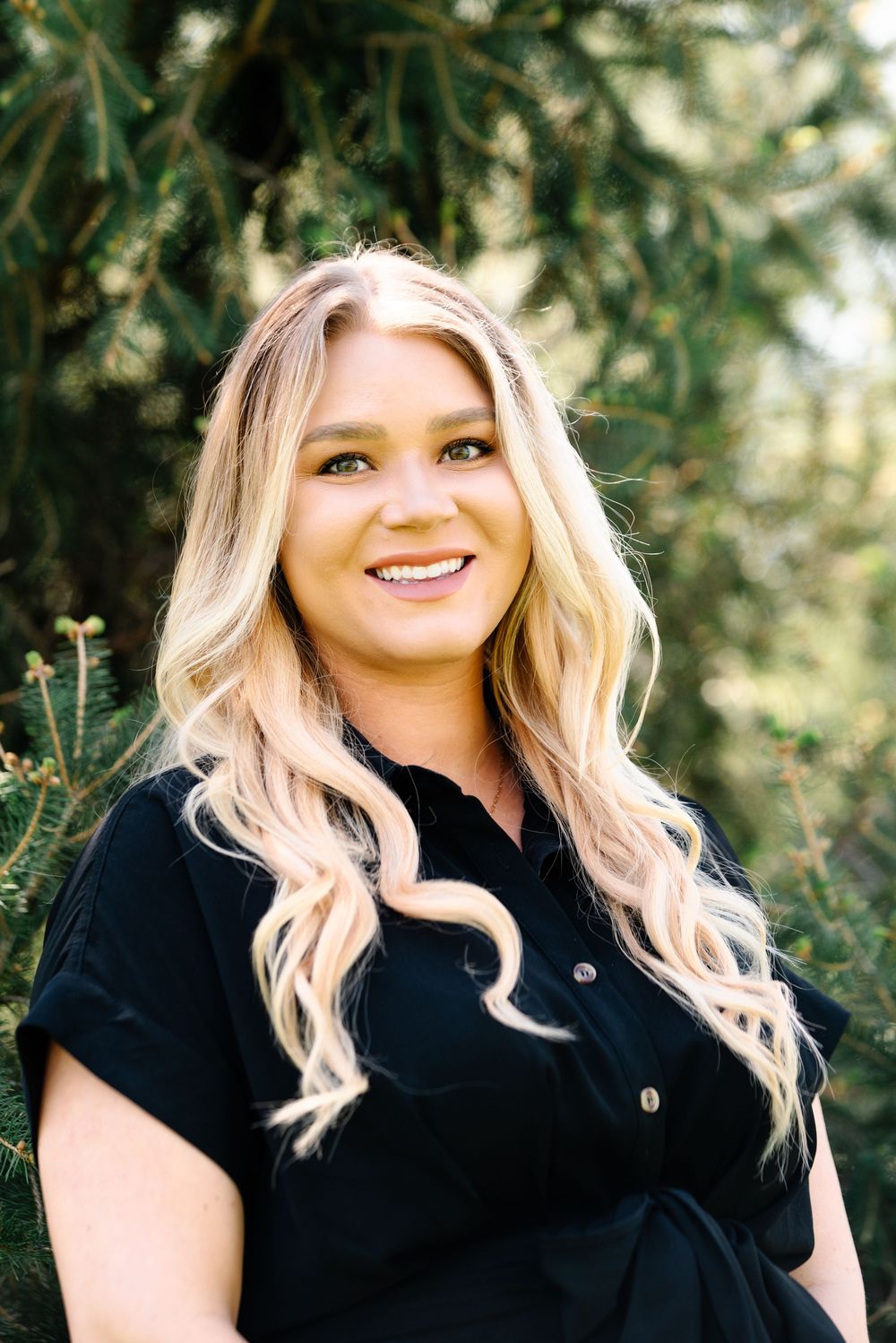 Smiling blonde woman in black shirt poses outdoors among green trees in a professional portrait photo.