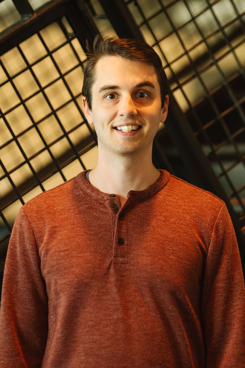 Smiling young man in a rust-colored henley shirt posing indoors with a modern glass ceiling background.
