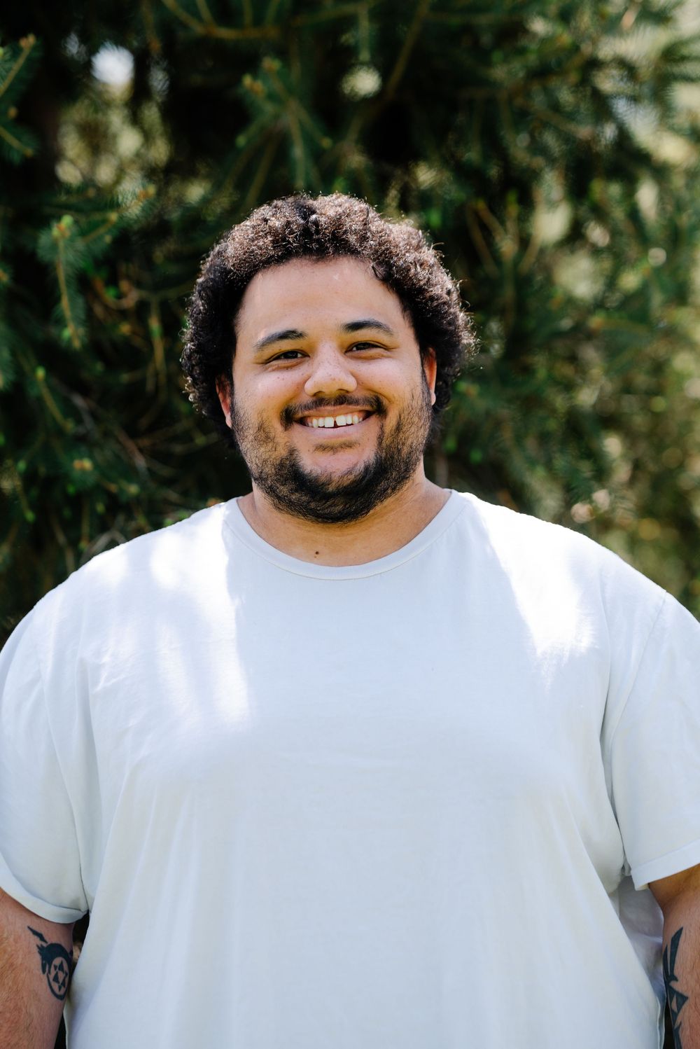 Smiling young man with curly hair and beard wearing a white t-shirt, standing outdoors with green trees behind him.