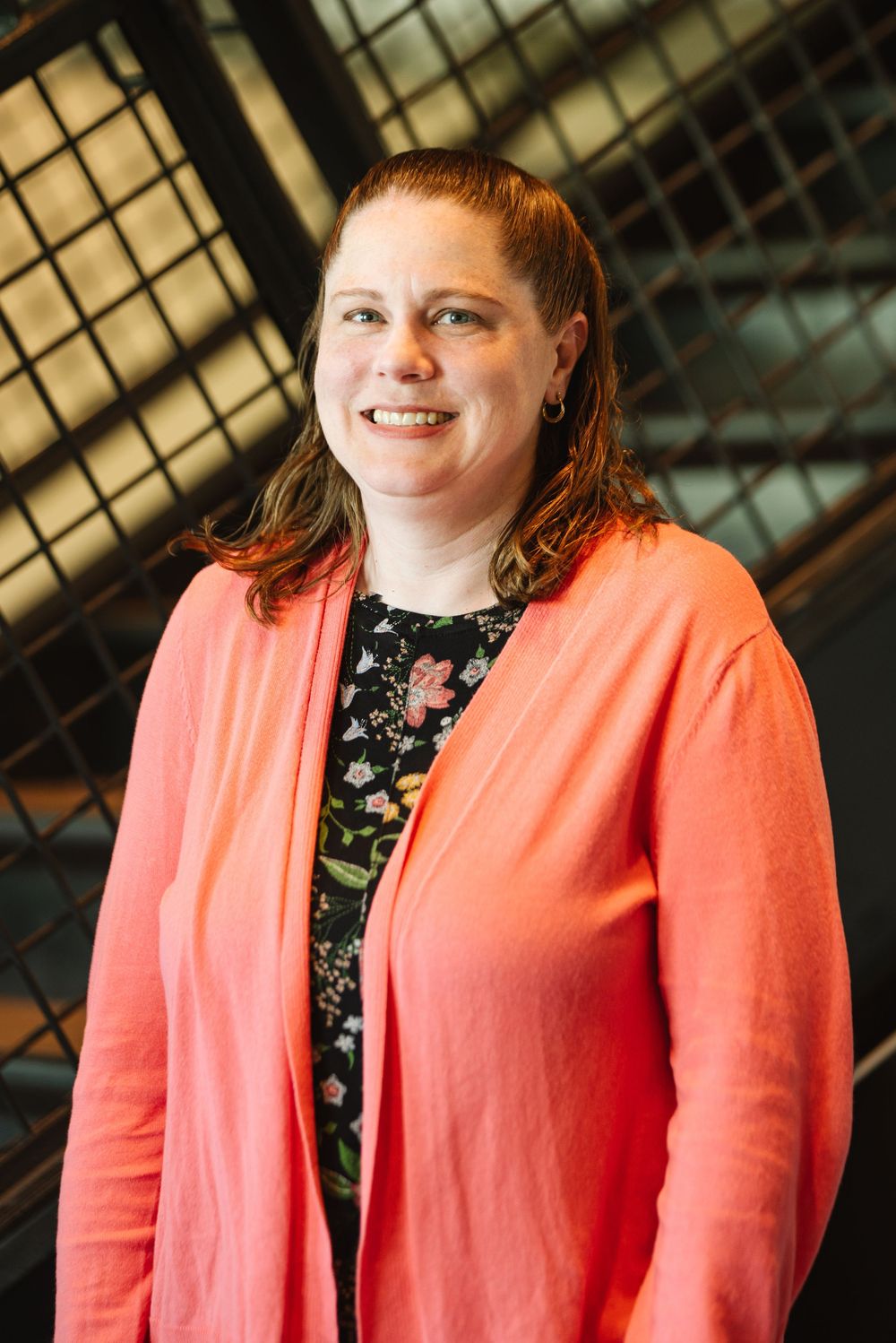 Smiling woman with red hair wearing a coral cardigan over a floral top, posing indoors with a metal grid background.
