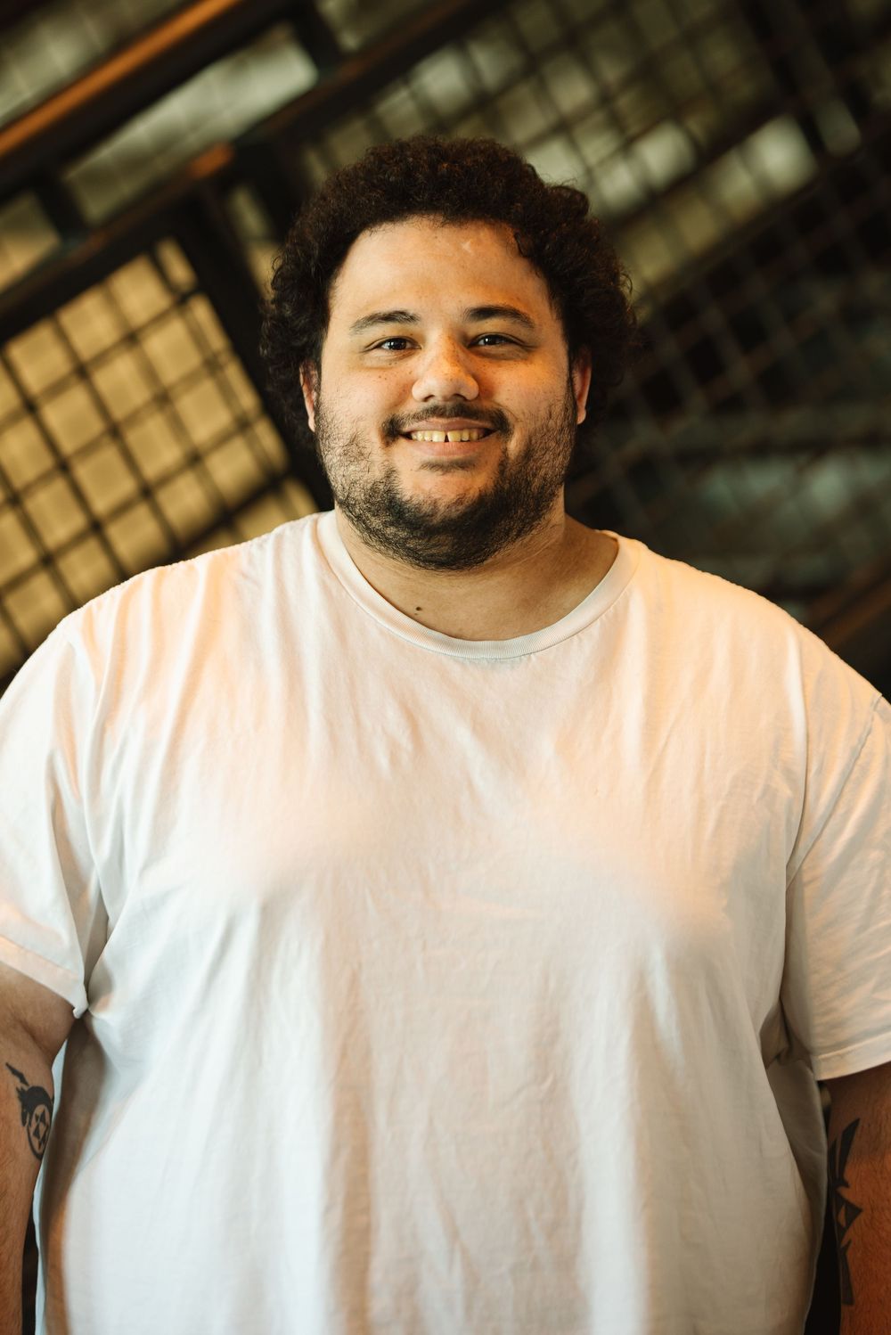 Smiling young man with curly hair and beard wearing a white t-shirt, showing tattoos on arms.