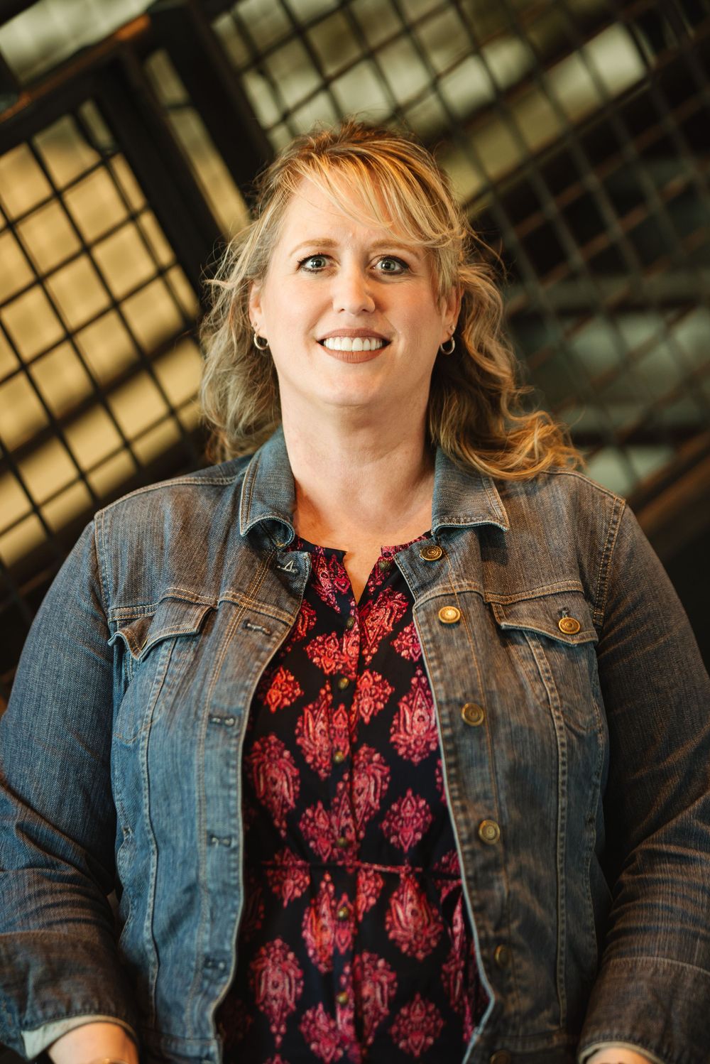Smiling woman with blonde hair wearing a denim jacket and floral top, posing indoors with a warm expression.