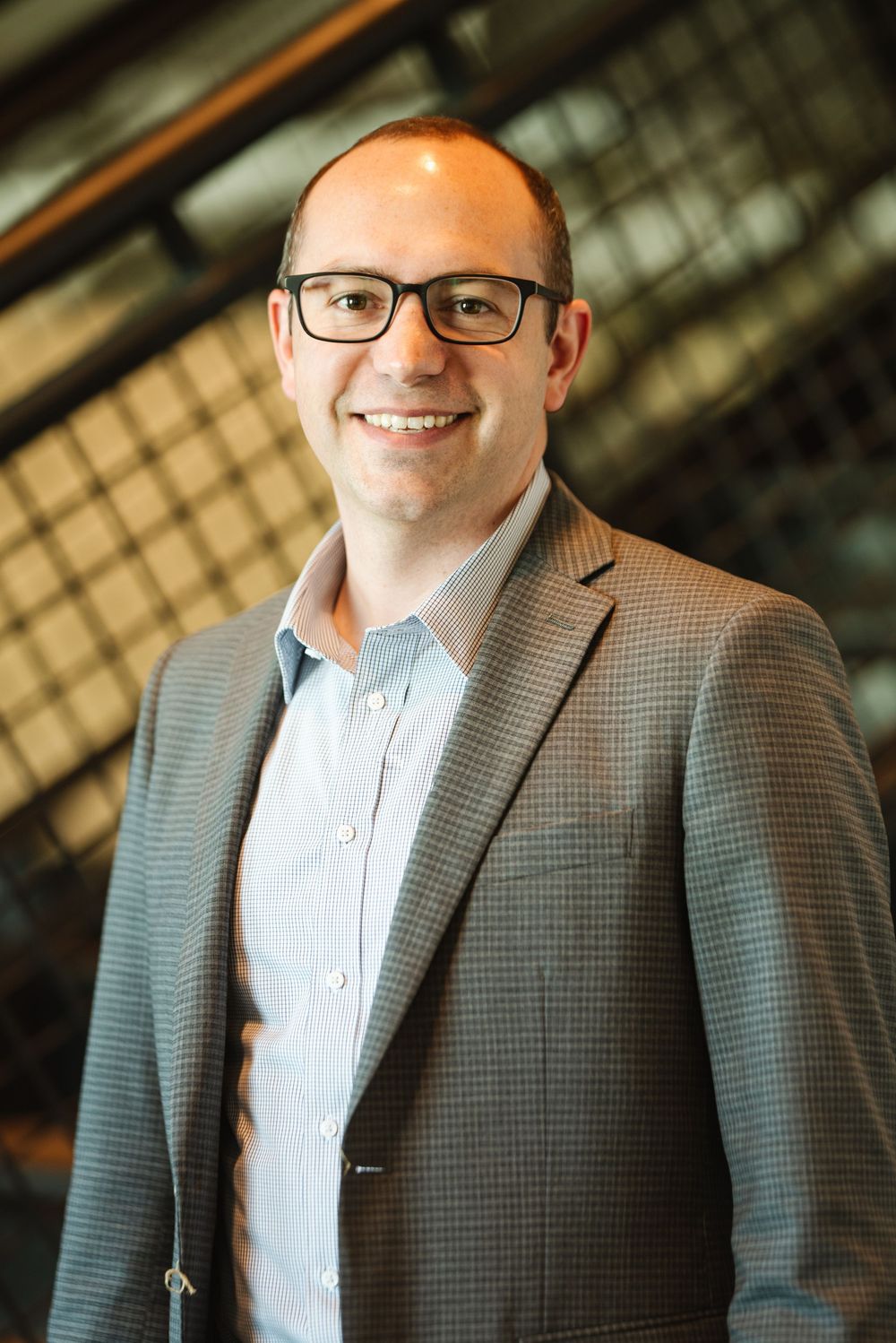 Professional man in gray blazer and glasses smiling confidently in a modern office building.