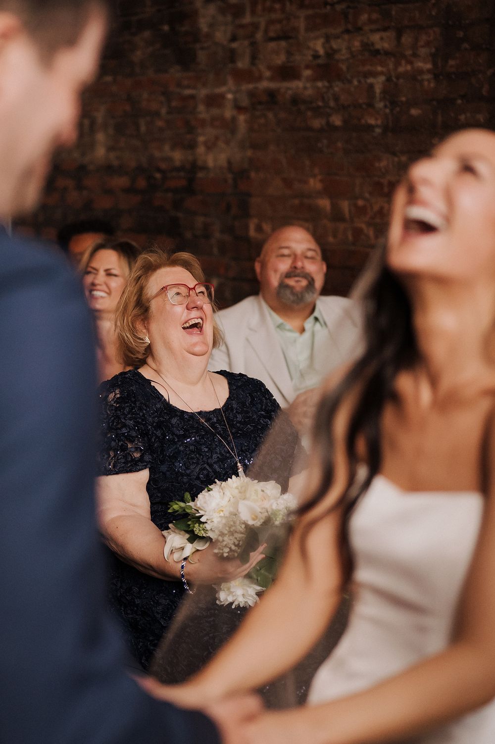 Joyful wedding guests laughing during ceremony, bride holding white bouquet, brick wall background.