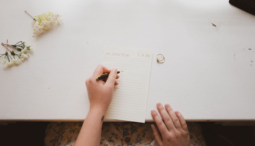 Person writing on a lined notepad on a white desk with flowers and a ring nearby.