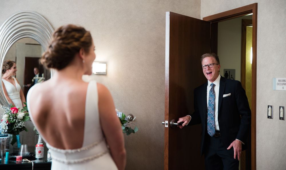 Father sees bride in wedding dress for first time, reacting with joy and surprise in hotel room doorway.