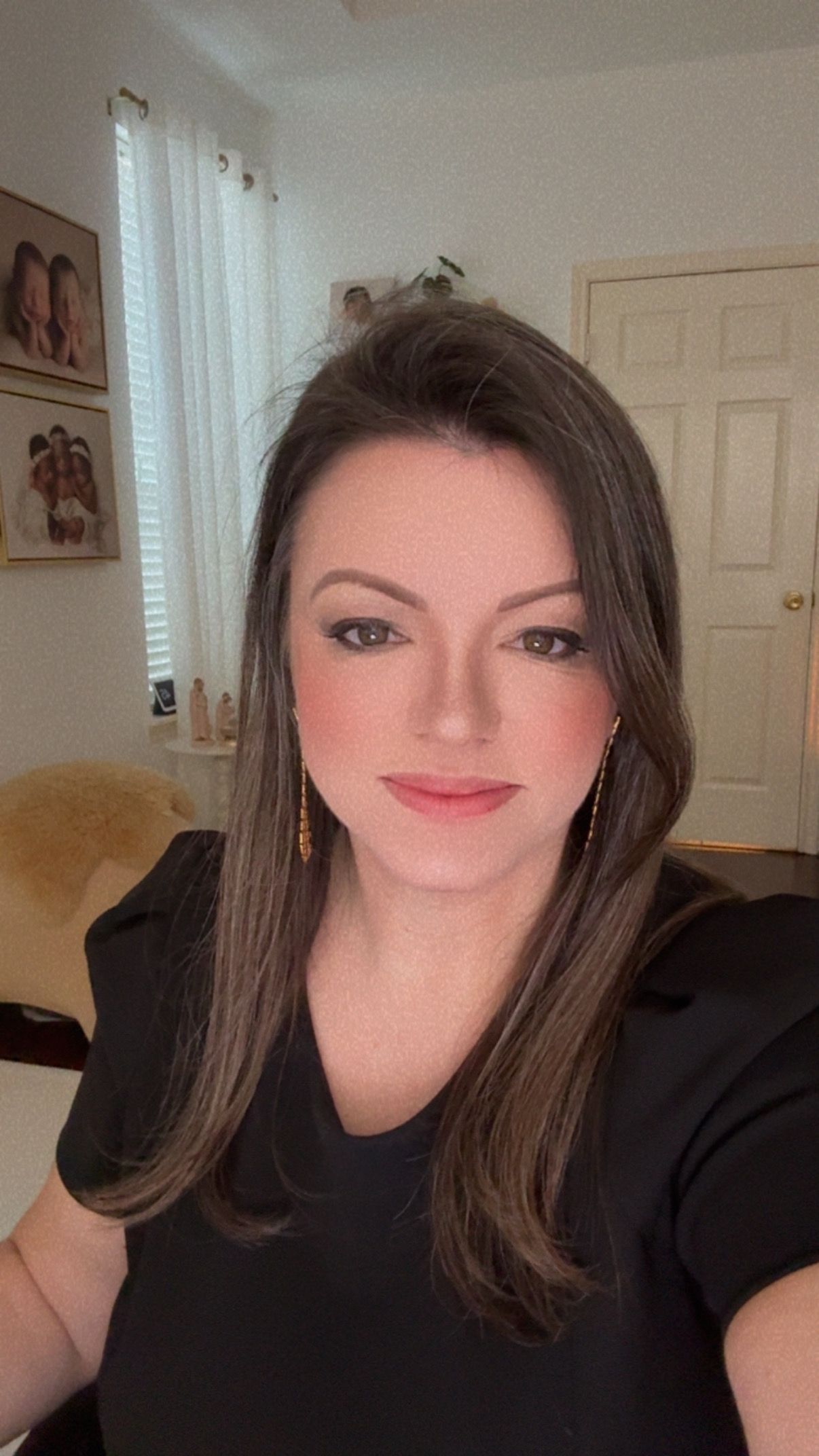A woman with dark brown hair wearing a black top and orange earrings takes a selfie in a well-lit room.