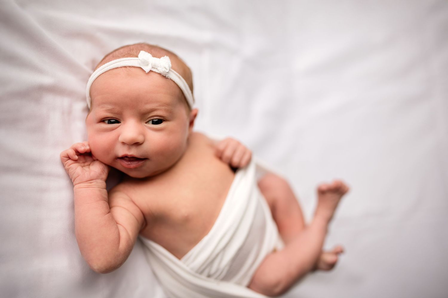 Newborn baby girl wrapped in white swaddle cloth wearing a white bow headband lying on white bedding.