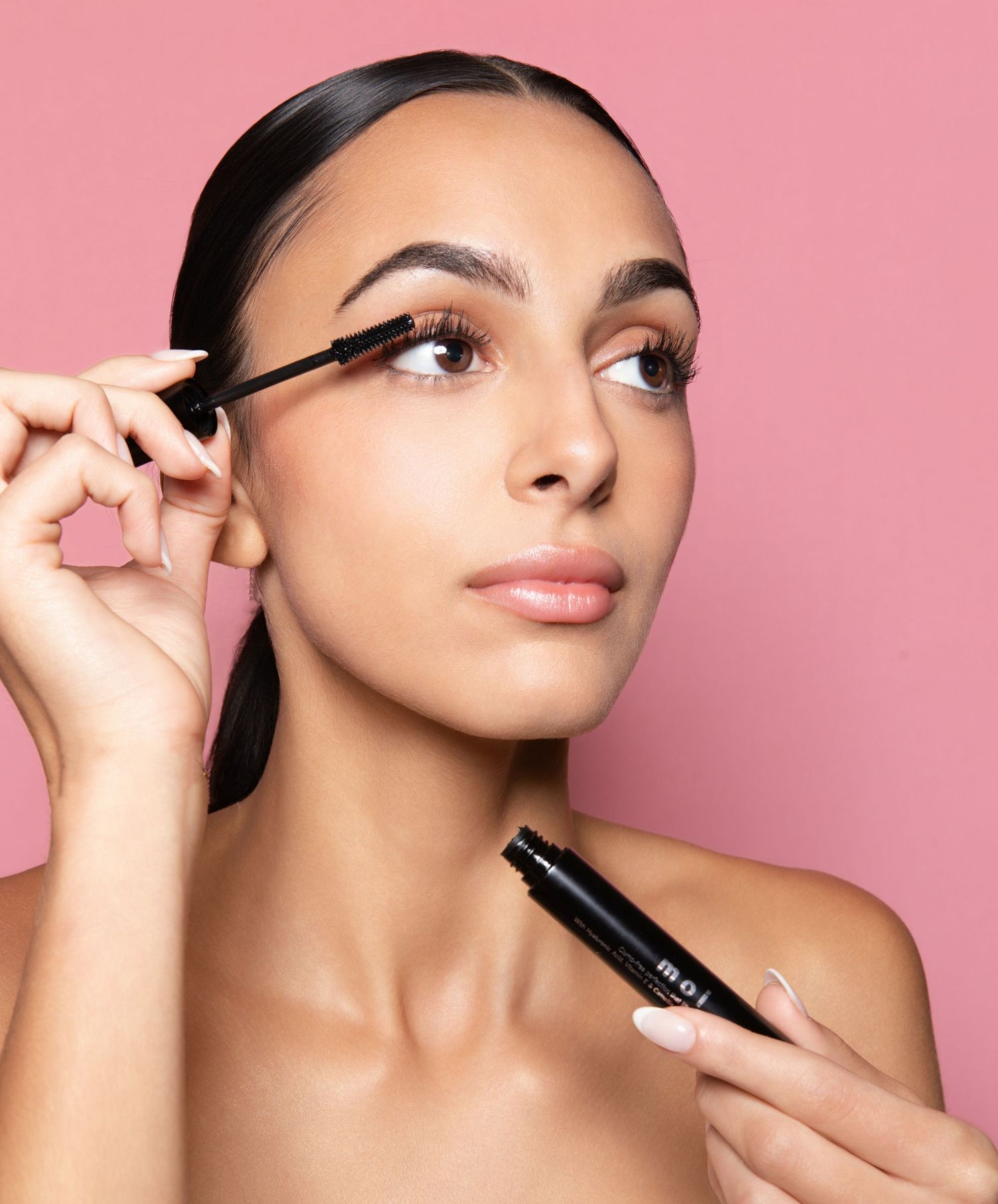 Woman applying black mascara with a wand, holding a mascara tube, against a pink background.