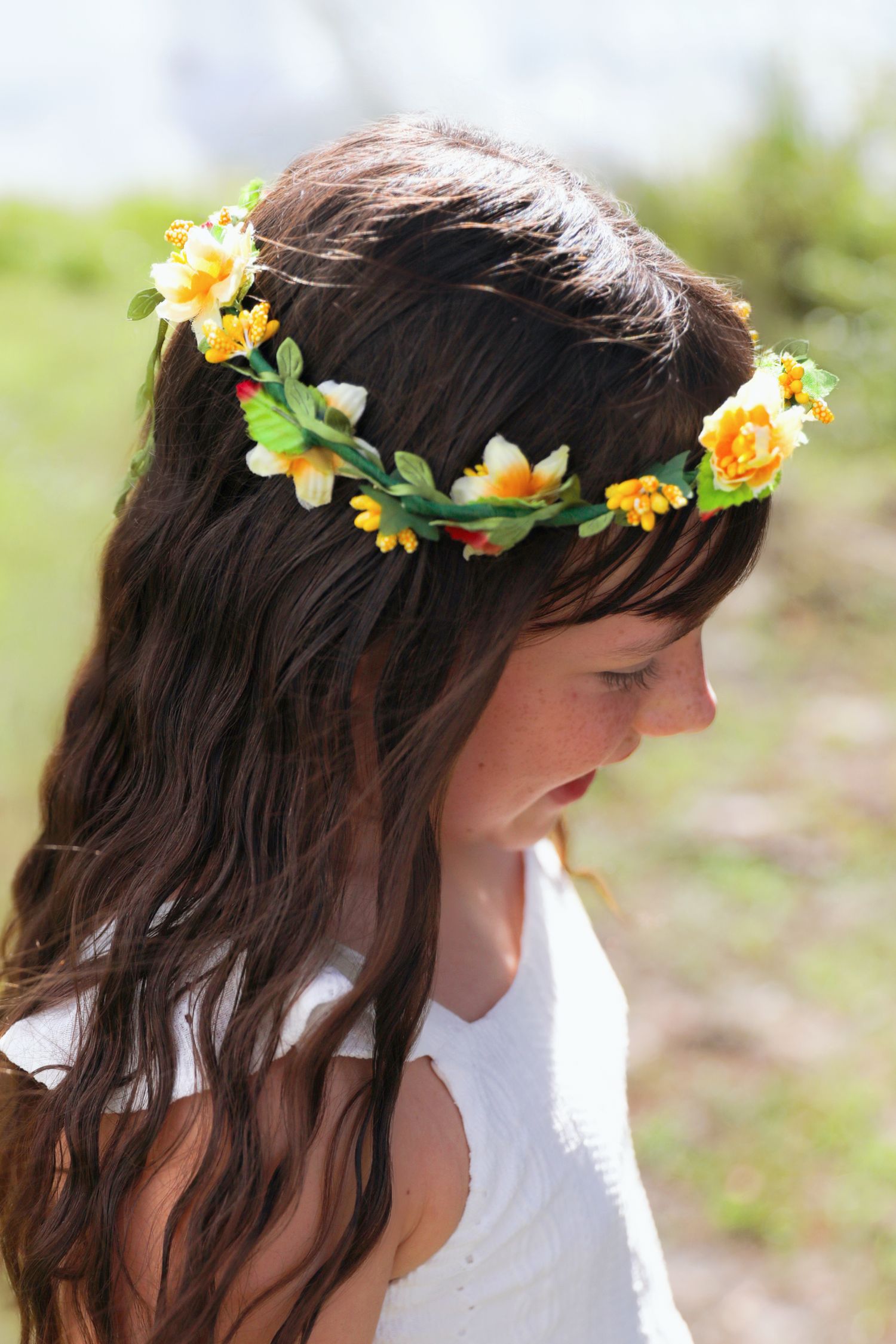 Young girl with long brown hair wearing a yellow and white flower crown outdoors in a sunny field.