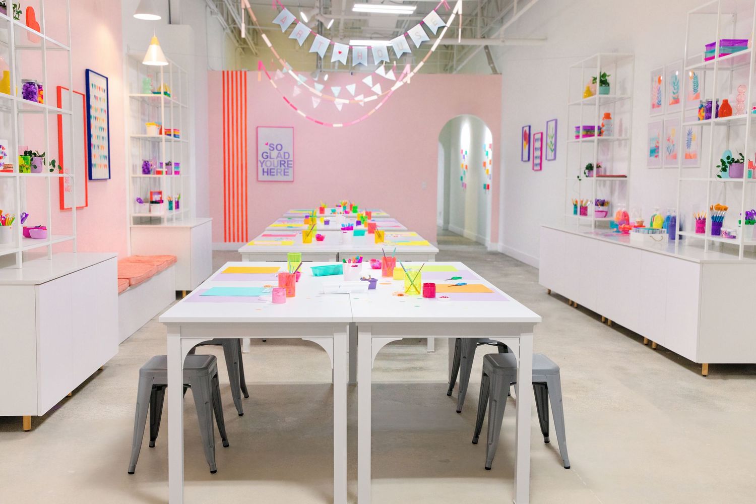 Bright and colorful craft studio with white tables, gray chairs, and shelves filled with colorful supplies.