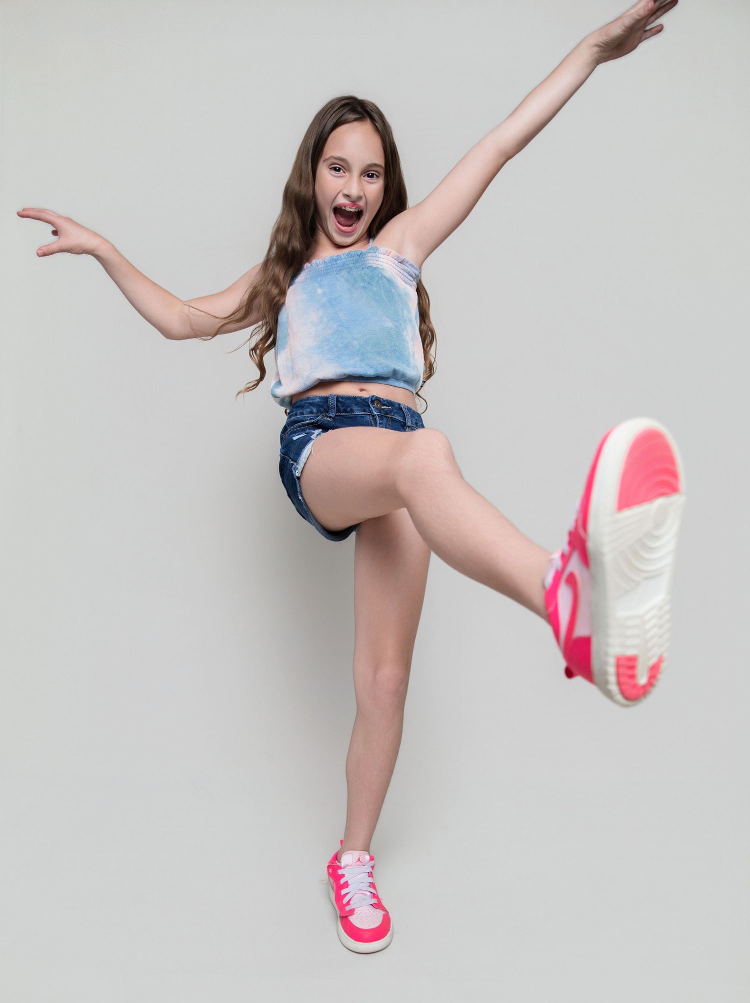 Excited young girl jumping with arms outstretched, wearing denim shorts, blue crop top, and pink sneakers.