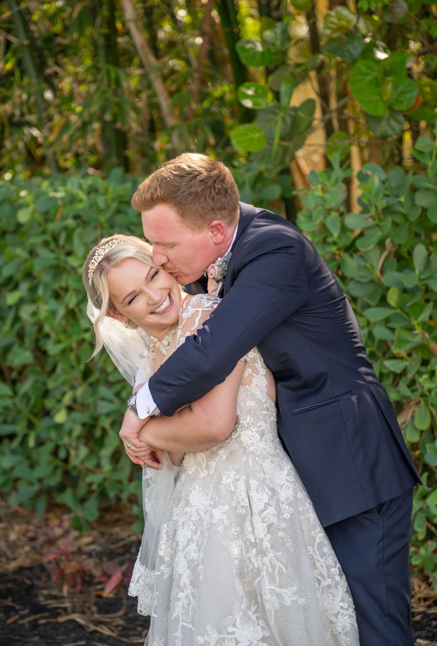 Bride in lace gown laughs as groom in navy suit hugs and kisses her cheek outdoors among lush greenery.
