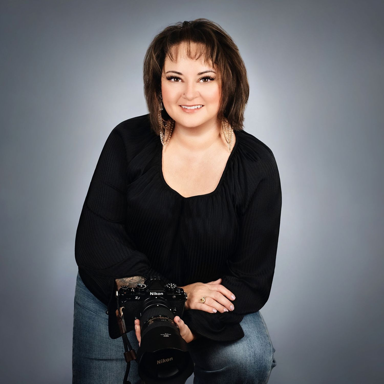 Woman in black top holding a Nikon camera, smiling confidently against a gray studio background.