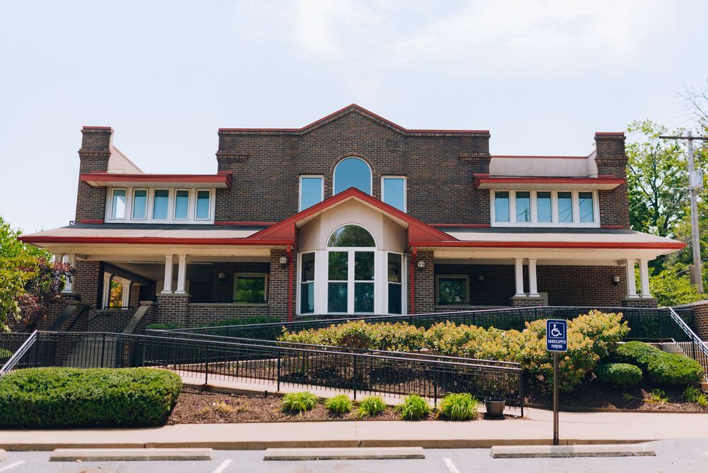 Brick building with red trim, arched entrance, ramps, and lush greenery under a clear sky.